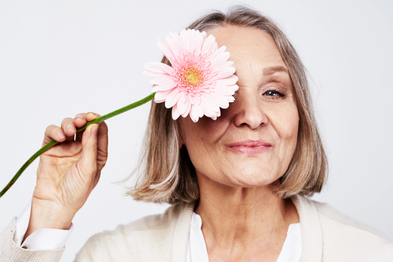 Äldre kvinna som håller en rosa blomma framför ena ögat
